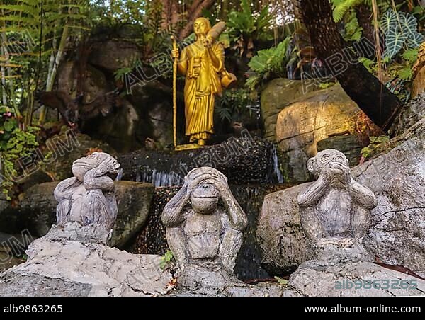 Buddhism symbols: gilded statue of Buddha as walking monk with Naga walking stick and umbrella and three wise monkeys statues sitting in foreground. Religion, diversity, inclusivity, prayer, worship.