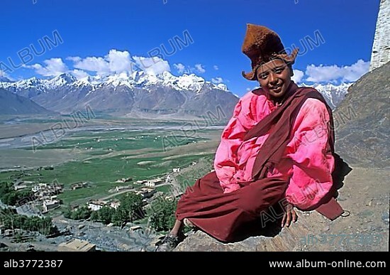 Young Buddhist monk wearing monk's robes sitting on a rock, in front of the small village of Karsha and the mountain ranges of the Himalayas, Karsha Monastery, Zanskar, Himalaya, North India, India, Asia.