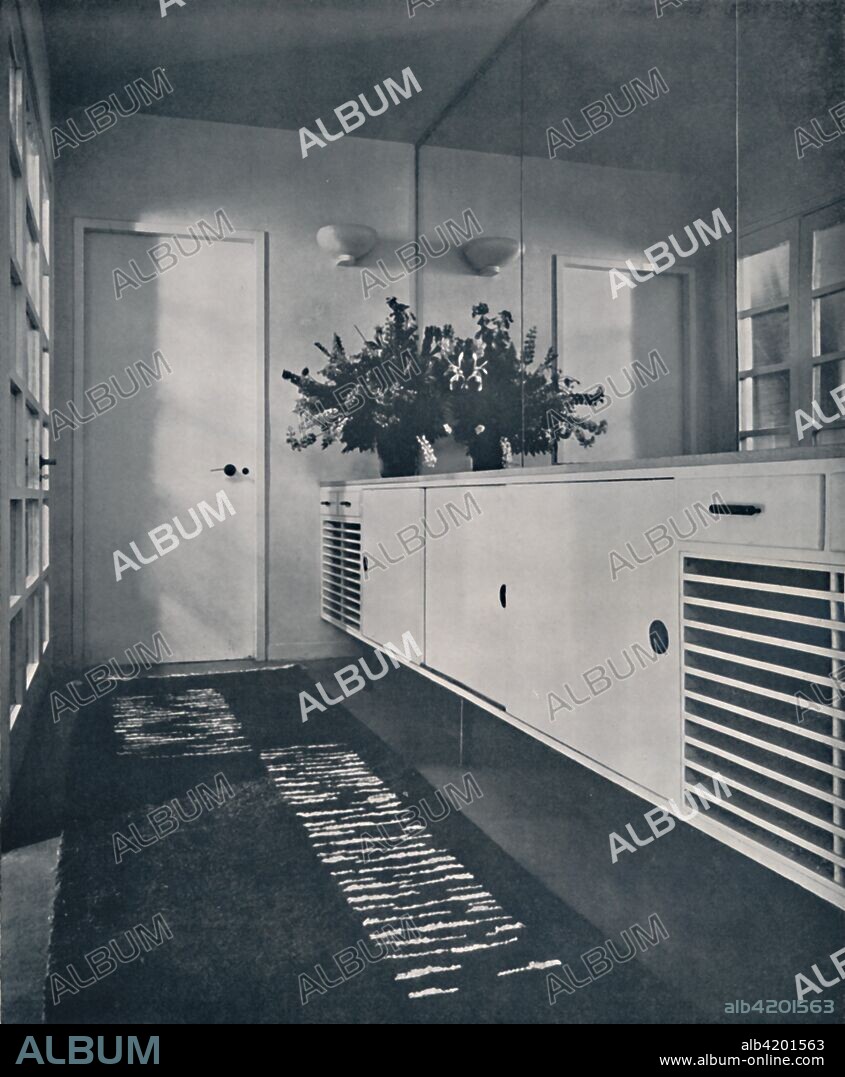 'Entrance lobby at Shrubs Wood, Chalfont St. Giles, England. Designed by Erich Mendelsohn and Serge Chermayeff, F.R.I.B.A.', 1936. Mirror-glass wall with chromium-plated holding bands, fitment incorporates cupboards and radiator-grilles, with waxed oak top. Floor of 'Ebernite' with hand-tufted rug. From Decorative Art 1936 - Year-Book of The Studio, by C.G. Holme. [The Studio Limited, London, 1936].