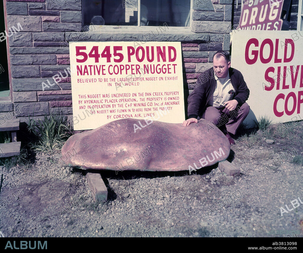 Historical photograph of a man posing with a large copper nugget. The sign behind the man reads: 5445 pound native copper nugget. Believed to be the largest copper nugget on exhibit in the world. This nugget was uncovered on the Dan Creek Property by hydraulic placer operation. The property is owned and in operation by the C + P Mining Co. of Anchorage.