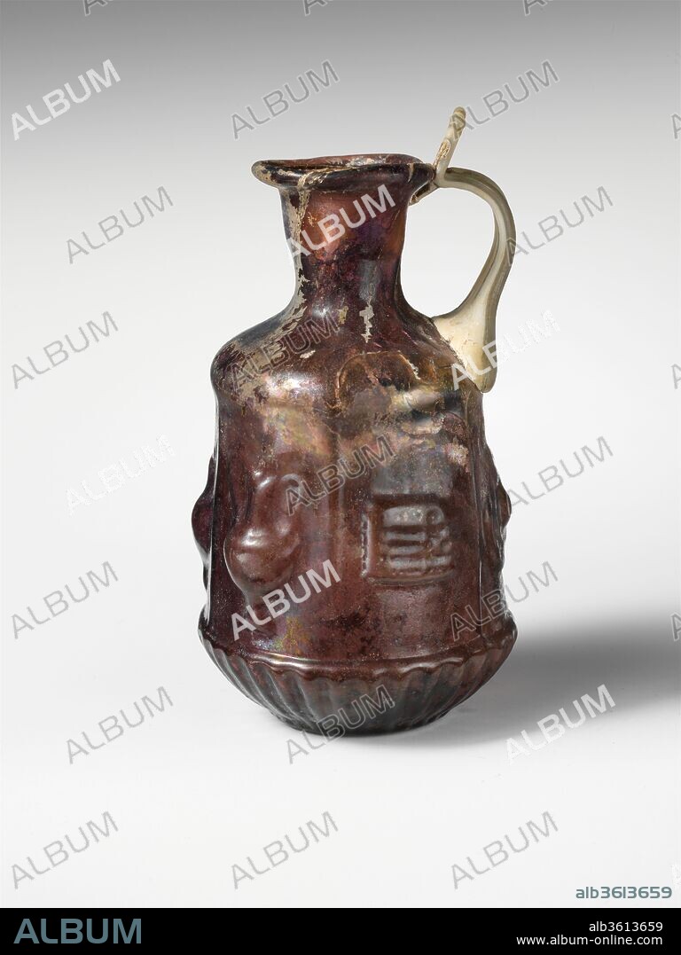 Glass hexagonal jug with Dionysiac symbols. Culture: Roman. Dimensions: H.: 3 7/8 in. (9.8 cm). Date: 1st century A.D..
Translucent deep purple, with handle in semi-opaque colorless with greenish tinge.
Tubular rim, folded out, round, and into flaring mouth; cylindrical neck, tapering downward; convex sloping shoulder; straight-sided hexagonal body, expanding downward; convex undercurve; flat circular bottom; strap handle attached in two large ribs to shoulder and top edge of body above panel with circular object, drawn up and out, then curved in, and pressed on to top of neck and underside of rim, with projecting flattened thumbrest above. Slight mis-alignment of vertical sections of mold on one side above cup section.
On shoulder, frieze of indistinct downturned tongues; on body, six rectangular panels, with vertical raised edges, containing objects associated with the Dionysiac cult: 1. crossed, double-ended thyrsi; 2. a footed jug with spout to left and high handle to right; 3. a footed amphora with high handles; 4. a syrinx (pan pipes) suspended horizontally from a vertical string; 5. a circular object with central protruding boss (a phiale mesomphalos or, perhaps, a cymbal); 6. a footed crater with high handles; around bottom, a frieze of thirty-one upturned, rounded tongues in raised relief; on bottom, raised circle around edge, a fine shallow circle surrounding a thicker and higher ring around a hollow central boss.
Broken, with several cracks and two holes, one in shoulder, the other in body below handle; handle streaked with black impurities; dulling, iridescence, and patches of whitish weathering on exterior, creamy brown weathering and iridescence on interior.