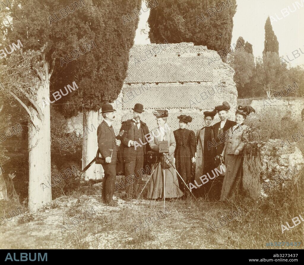 Group portrait of five women (one of whom is behind a camera) and two men for a ruin in Italy or France, Anonymous, c. 1885.