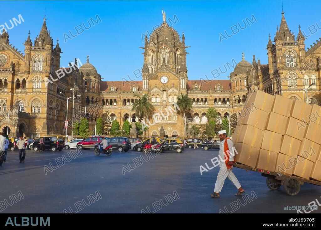 India: A man with fully laden hand cart in front of Chhatrapati Shivaji Maharaj Terminus, Mumbai, a UNESCO World Heritage Site and the city's busiest railway station. This impressive railway station, formerly named Victoria Terminus, was built between 1878 and 1887, the station is an example of Victorian Italian Gothic style and was modelled on St. Pancras Station in London. The roof has church-like little spires and an impressive, gothic-style central dome. The terminus was designed by Frederick William Stevens from an initial design by Axel Haig.