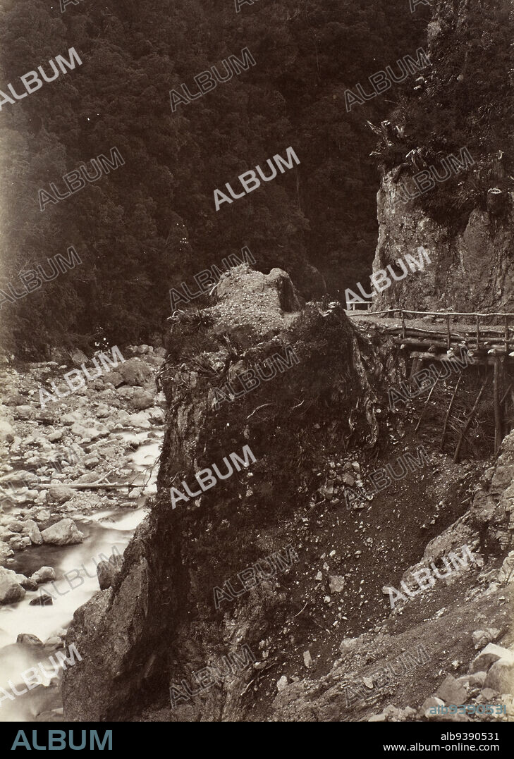 Otira Gorge, West Coast Road, Burton Brothers studio, photography studio, Dunedin, black-and-white photography, View looking down a section of Otira Gorge landscape. River waters can be seen along the facing left hand side of the image, and a built up road can be seen on the facing right hand side. The background hillside is clad in native bush and trees,while the riverbank landscape, at ground level, is covered by a combination of rocks and stones.