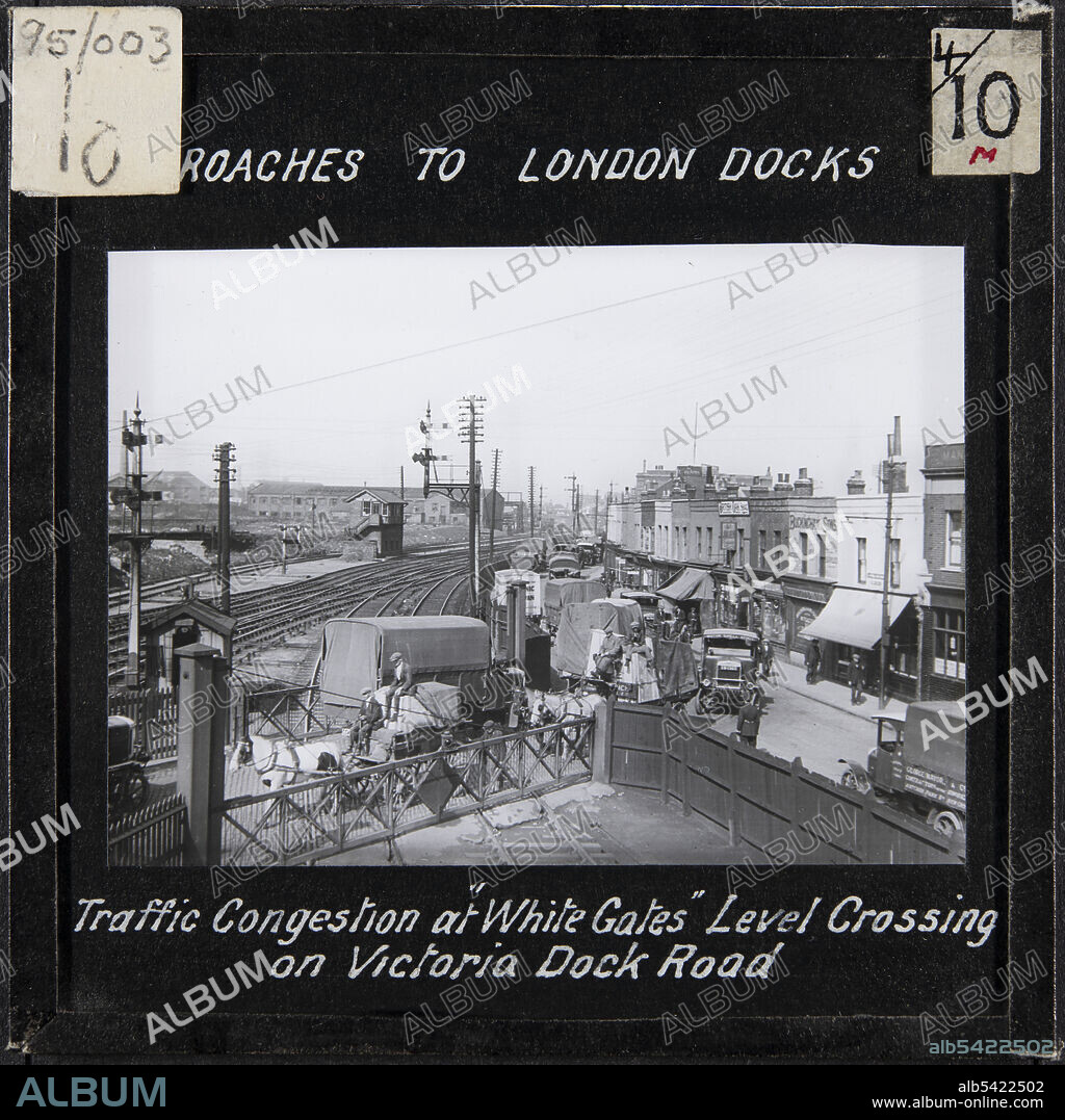White Gates Level Crossing, Canning Town, Newham, Greater London Authority, 1920s. Vans and horse-drawn carts on the White Gates Level Crossing, and traffic queuing on Victoria Dock Road alongside the North Woolwich RailwayThe caption on this slide reads: "Approaches to London Docks. Traffic Congestion at "White Gates" Level Crossing on Victoria Dock Road."The White Gates Level Crossing provided vehicular access from Victoria Dock Road to the Royal Victoria Dock and Silvertown, crossing the North Woolwich Railway. In 1922 the average number of vehicles crossing White Gates in 24 hours was 2,852, an increase from 2,173 in 1904 and 2,496 in 1913. Due to closure of the gates for trains, road traffic was stopped for over 9 hours each day. The level crossing was therefore one of a number of bottle necks on the approach to the docks, which also included the narrow swing bridge at the west entrance of Royal Victoria Dock. It was not until the Royal Victoria and other Dock Approaches (Improvement) Act was passed in 1929 that work began on a new road scheme in the area.