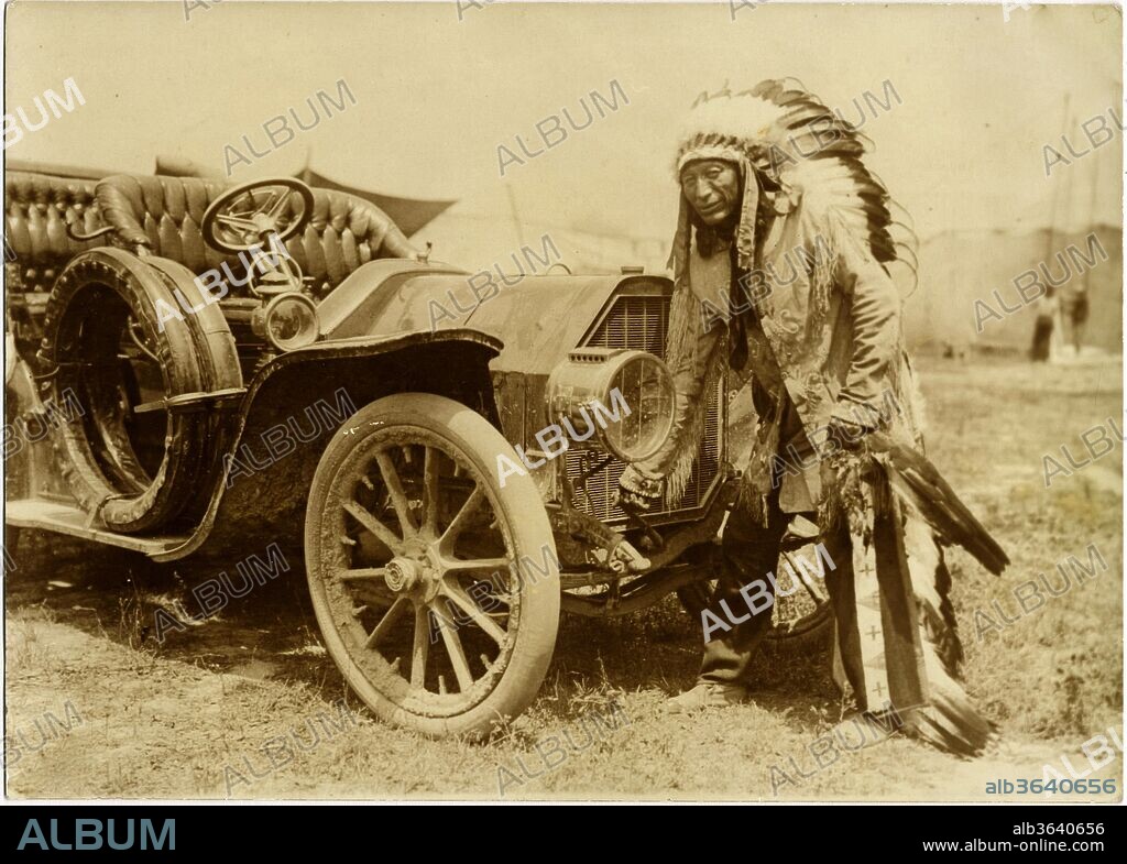 Native American Indian With Full Regalia And Headdress. From Horse To Automobile - Circa 1915. The Native American Indian Chief In Extensive Ceremonial Dress Cranking A Large, Early Automobile Must Have Seen The Humor In This Photograph, As He Has A Slight Smile. The Irony Of The Transition That The United States Had Undergone During The Industrial Revolution, Turning The Native American Indian Into An Operator Of Automobiles And Not His Horse., 1915.