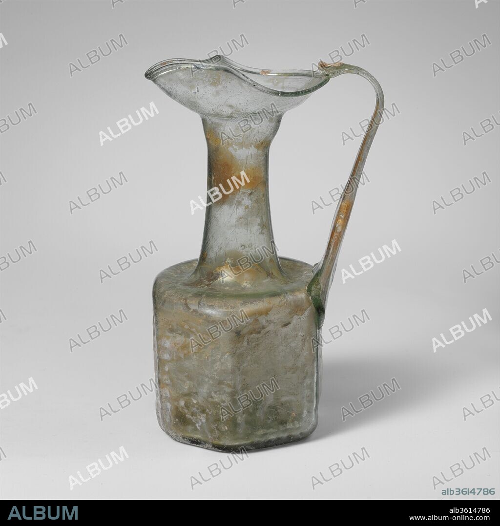 Glass hexagonal jug with Jewish symbols. Culture: Roman, Palestinian. Dimensions: Height  5 11/16in. (14.4cm); diameter  2 3/8in. (6.1cm). Date: late 6th-early 7th century A.D..
Translucent light green; handle in same color.
Mouth and neck free blown. Rounded, partially tubular rim, folded out and down; broad trefoil mouth with narrowed spout; cylindrical neck with bulge at top, then expanding downwards; concave shoulder with rounded outer edge; hexagonal body with concave sides; pushed-in bottom with small central kick and circular pontil scar; broad strap handle applied in uneven pad to top of body and edge of shoulder, drawn up and outwards in a straight line, then curved in and trailed off on rim opposite spout with slight upward trail folded back on top of handle.
On the body, six rectangular panels with impressed decoration, each of a different design bordered by lines of recessed dots. They comprise a five-branched menorah on a tripod base, a double lozenge with diamond-shaped central boss, and various vegetal motifs, including a stylized palm tree.
Complete, but crack across middle of handle; pinprick and elongated bubbles, with some black impurities in handle and rim; dulling, slight pitting, and faint whitish iridescent weathering, with patches of thicker creamy brown weathering on interior of neck and handle.
Such hexagonal and octagonal jars or jugs were mass-produced in molds for the pilgrim trade in the Holy Land. This example, found in Cyprus, bears witness to the important place the island had on the sea route that brought pilgrims to Jerusalem.