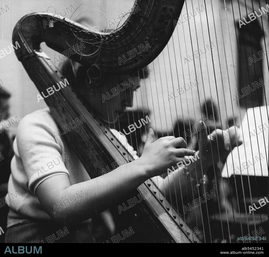 Student Of The Harp -- Playing With The School Orchestra: Sheila Bromberg photographed during a performance of the Guildhall School Orchestra. - The loose ends of the harp strings are not the outcome of untidiness; harpist's use the extra length for repairs. January 31, 1951. (Photo by Tom L. Blau, Camera Press).