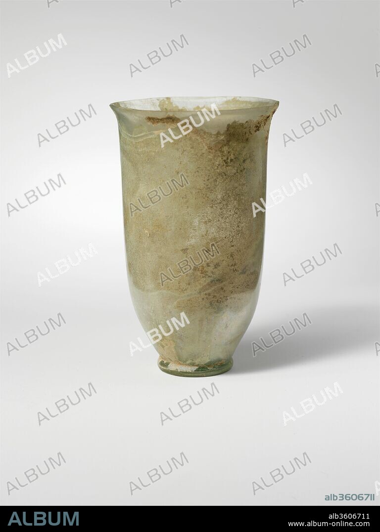 Glass beaker. Culture: Roman. Dimensions: H.: 3 5/8 in. (9.2 cm). Date: 1st century A.D..
Colorless with pale blue green tinge.
Knocked-off, uneven rim; slightly bulging collar below rim; narrow, cylindrical body with straight side, then tapering in to folded foot ring; small bottom with low central kick.
Intact, except for small chip in rim; some bubbles and blowing striations; iridescence and creamy weathering, with thick soil encrustation around bottom of interior.