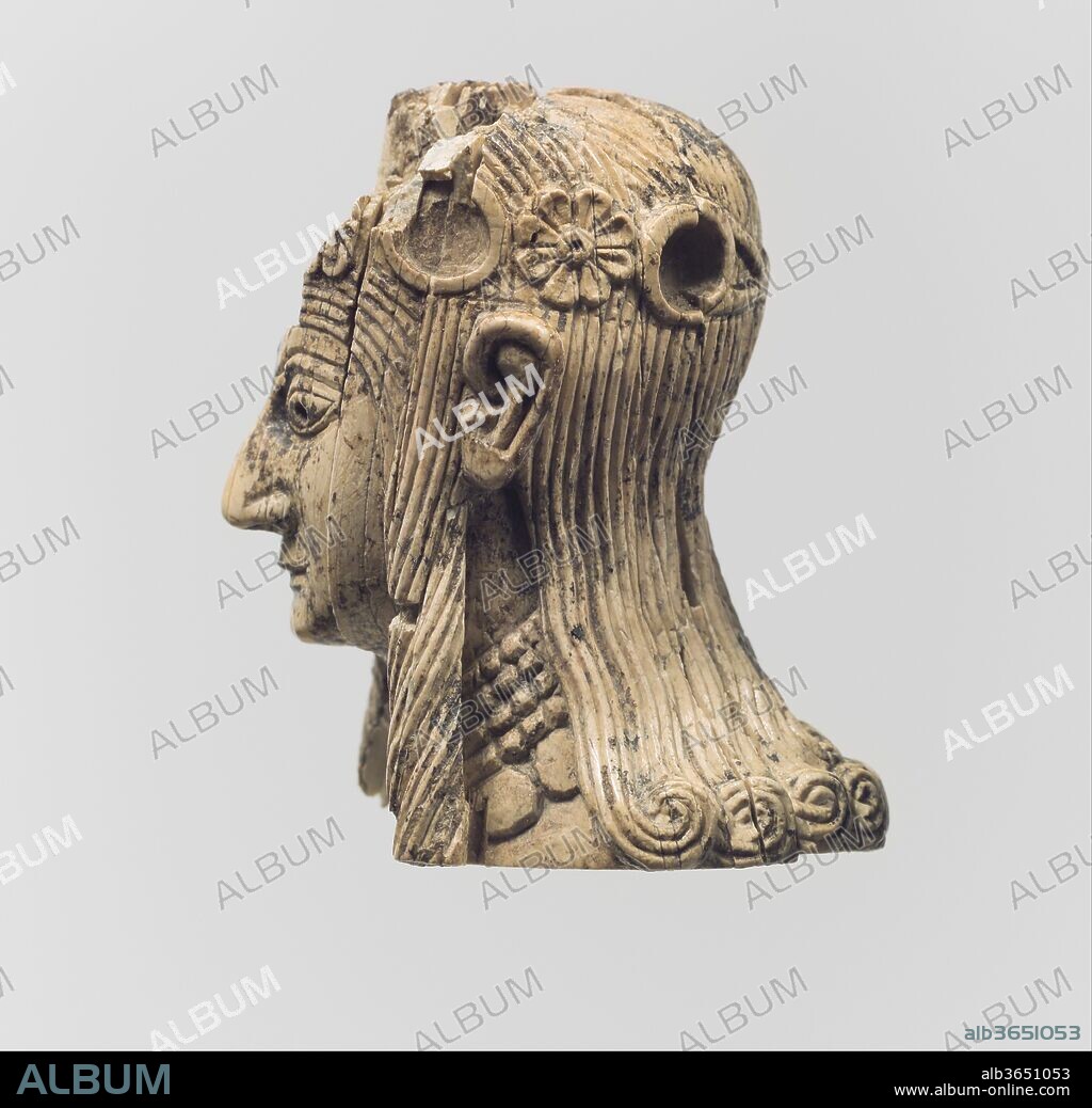 Head of a female figure. Culture: Assyrian. Dimensions: 1.97 x 1.22 in. (5 x 3.1 cm). Date: ca. 8th-7th century B.C..
This female head, carved in the round, was found in the Burnt Palace at Nimrud and may have originally been part of a composite statuette made of various materials and overlaid with gold foil. A square mortise cut into the bottom of this piece suggests that a tenon would have secured it to a now lost body. Part of the head has been damaged, but several features that remain, including the large, originally inlaid eyes, prominent ears, hooked nose, small mouth, and receding chin, are characteristic of North Syrian ivories. The hair falls in long individual locks, crowned by a diadem of rosettes and inlaid discs (the original colored glass or semiprecious stone inlays are now lost) tied at the back of the head. The choker-style necklace with pendants worn by this female figure is similar in form to those worn by the female figures depicted on an ivory handle also found in the Burnt Palace and in the Metropolitan Museum's collection (MMA 52.23.2).
Built by the Assyrian king Ashurnasirpal II, the palaces and storerooms of Nimrud housed thousands of pieces of carved ivory. Most of the ivories served as furniture inlays or small precious objects such as boxes. While some of them were carved in the same style as the large Assyrian reliefs lining the walls of the Northwest Palace, the majority of the ivories display images and styles related to the arts of North Syria and the Phoenician city-states. Phoenician style ivories are distinguished by their use of imagery related to Egyptian art, such as sphinxes and figures wearing pharaonic crowns, and the use of elaborate carving techniques such as openwork and colored glass inlay. North Syrian style ivories tend to depict stockier figures in more dynamic compositions, carved as solid plaques with fewer added decorative elements. However, some pieces do not fit easily into any of these three styles. Most of the ivories were probably collected by the Assyrian kings as tribute from vassal states, and as booty from conquered enemies, while some may have been manufactured in workshops at Nimrud. The ivory tusks that provided the raw material for these objects were almost certainly from African elephants, imported from lands south of Egypt, although elephants did inhabit several river valleys in Syria until they were hunted to extinction by the end of the eighth century B.C.