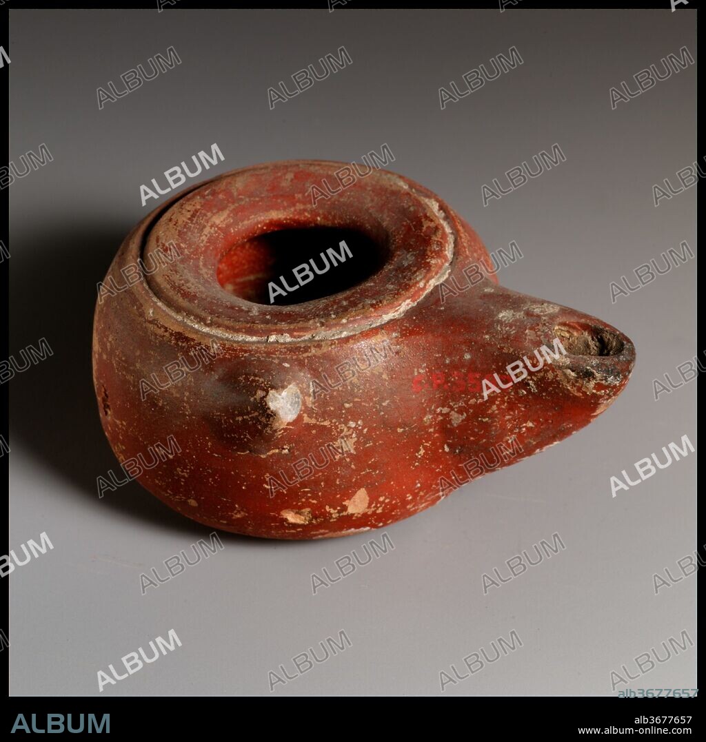 Terracotta oil lamp. Culture: Greek. Dimensions: Overall: 1 1/2 x 3 1/4 in. (3.8 x 8.3 cm).
Wheel-made body, with applied long nozzle. Large central filling hole, surrounded by an inward-sloping narrow shoulder and a deep, incised circle; convex-curving side to cylindrical body and a single projecting knob at top edge of left side. Raised base ring and concave base. Red slip.
Intact. Part of base ring pushed in before firing. Heavy, thick base.