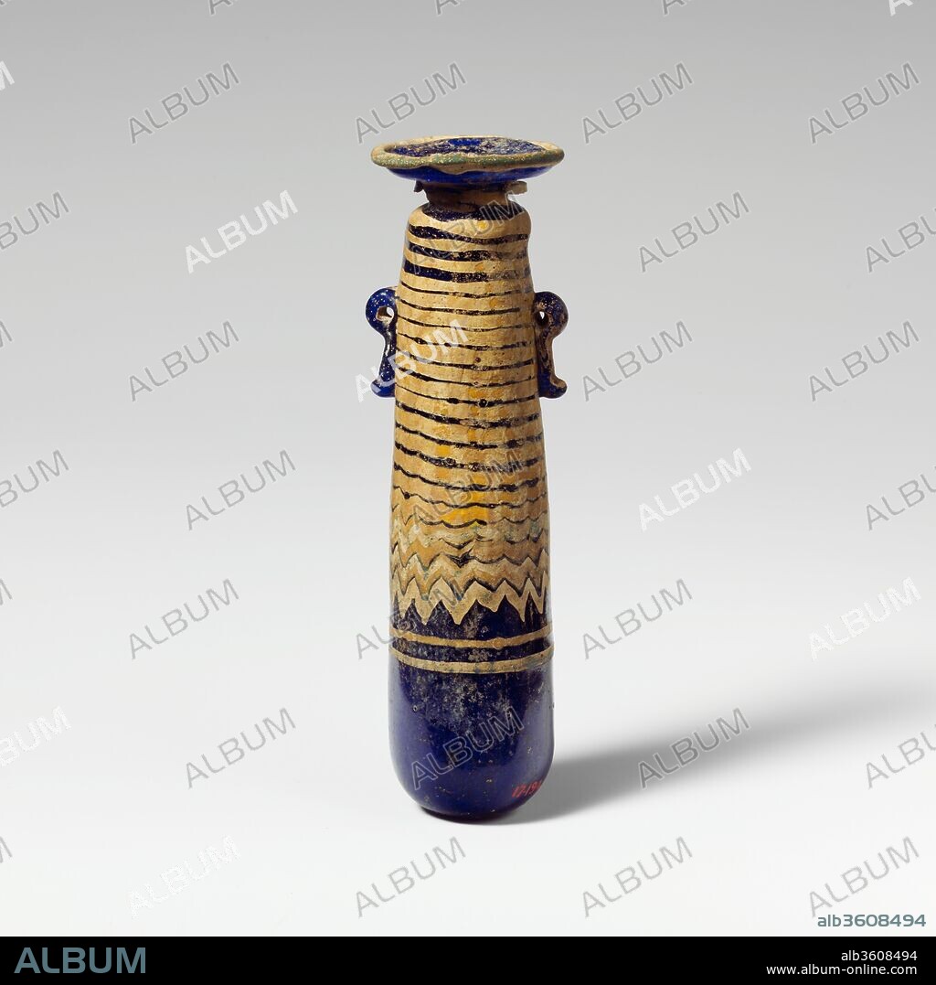 Glass alabastron (perfume bottle). Culture: Greek, Eastern Mediterranean. Dimensions: H.: 4 1/4 in. (10.8 cm). Date: late 6th-5th century B.C..
Translucent cobalt blue, with handles in same color; trails in opaque yellow and opaque turquoise blue.
Broad, inward sloping rim-disk, with radiating tool marks on upper surface and uneven edge around mouth; short cylindrical neck with downward taper; narrow rounded shoulder; straight-sided cylindrical body, tapering upwards; convex bottom; two vertical ring handles with knobbed tails, applied over trail decoration.
Turquoise blue trail attached at edge of rim-disk; a yellow trail applied on underside of rim-disk and wound down in a spiral to middle of body, then tooled into a close-set zigzag pattern; a turquoise blue trail is added, mingling with yellow; immediately below zigzag, another yellow trail and another turquoise blue trail are wound horizontally once around body.
Intact; dulling and milky iridescent weathering.