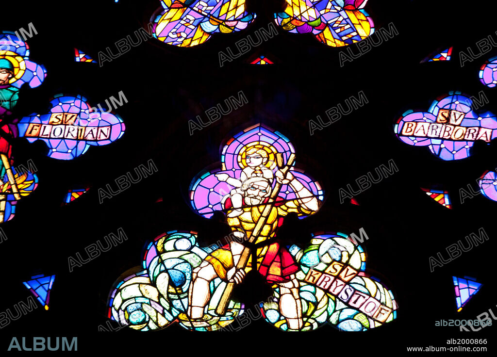 Prague, St. Vitus Cathedral, Thunov Chapel, Stained Glass Window, Psalm 126:5. Stained Glass window in the Thunov Chapel, in the St Vitus Cathedral, Prague, Czech Republic. The window was designed by Frantisek Kysela 1929 - 1934, depicting scenes according to Psalms 126:5: "They that sow in tears shall reap in joy". The window portrays natural disasters such as floods, hail storms, fire, death, injury and old age, coupled with biblical quotes. Seen is a close-up of the crest of the window, with St Krystof holding a wooden cane and bearing Baby Jesus in the center, flanked by St Florian and St Barbara (not seen in the photo).