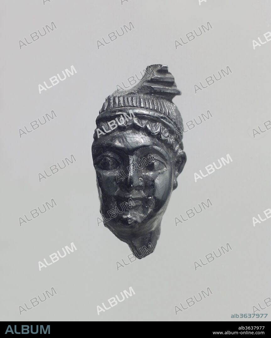 Head. Culture: Assyrian. Dimensions: 1.3 x 0.71 x 0.59 in. (3.3 x 1.8 x 1.5 cm). Date: ca. 8th-7th century B.C..
This carved head was probably blackened through exposure to fire when the palace complexes at Nimrud were sacked during the final defeat of Assyria at the end of the seventh century B.C. Originally, this piece may have been part of a composite statuette made of various materials and overlaid with gold foil. The figure is depicted with fleshy cheeks, large eyes and nose, and a small mouth. The hair appears as a row of curls along the forehead, held back by a plain fillet. A striated pattern and fragmentary additional element at top may represent hair topped by a headdress, although this is an unusual image not paralleled by other headdresses known from this period. Alternatively, the figure may be represented as a caryatid, a column in the form of a human figure, and the fragmentary element at top may be a column capital. As the head is beardless, it could represent either a male foreigner, a woman, or a eunuch. Eunuchs, who were castrated men, served in many high-level positions in the Assyrian court and are frequently represented on the stone reliefs that decorate the walls of the Northwest Palace at Nimrud. 
Built by the Assyrian king Ashurnasirpal II, the palaces and storerooms of Nimrud housed thousands of pieces of carved ivory. Most of the ivories served as furniture inlays or small precious objects such as boxes. While some of them were carved in the same style as the large Assyrian reliefs lining the walls of the Northwest Palace, the majority of the ivories display images and styles related to the arts of North Syria and the Phoenician city-states. Phoenician style ivories are distinguished by their use of imagery related to Egyptian art, such as sphinxes and figures wearing pharaonic crowns, and the use of elaborate carving techniques such as openwork and colored glass inlay. North Syrian style ivories tend to depict stockier figures in more dynamic compositions, carved as solid plaques with fewer added decorative elements. However, some pieces do not fit easily into any of these three styles. Most of the ivories were probably collected by the Assyrian kings as tribute from vassal states, and as booty from conquered enemies, while some may have been manufactured in workshops at Nimrud. The ivory tusks that provided the raw material for these objects were almost certainly from African elephants, imported from lands south of Egypt, although elephants did inhabit several river valleys in Syria until they were hunted to extinction by the end of the eighth century B.C.