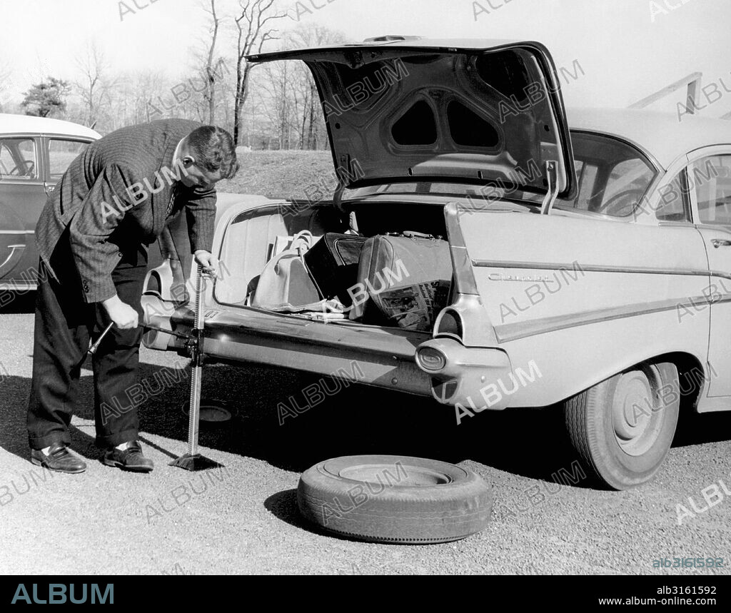 United States: 1958 A man jacking up his 1957 Chevrolet to change a rear tire on it.